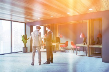 İki genç iş adamı modern açık uzay ofisinde gri ve ahşap duvarları, fayanslı zemini ve kırmızı sandalyeli sıralı bilgisayar masaları olan belgeleri tartışıyor. Tonu ayarlanmış resim