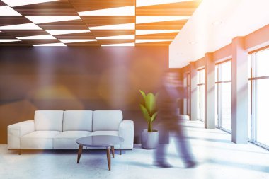 Bulanık genç işadamı panoramik ofis bekleme odasında gri duvarları, fayanslı zemini ve kahve masası olan rahat beyaz koltuğu ile yürüyor. Tonu ayarlanmış resim