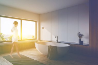 Beyaz ve ahşap duvarları, ahşap zemini, rahat küveti ve tropikal manzaralı penceresi olan geniş güneşli banyoda yürüyen bulanık genç kadın. Tonu ayarlanmış resim