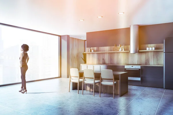 Beautiful young African American woman standing in panoramic kitchen with grey and wooden walls, tiled floor, gray countertops and fridge and bar counter with chairs. Toned image