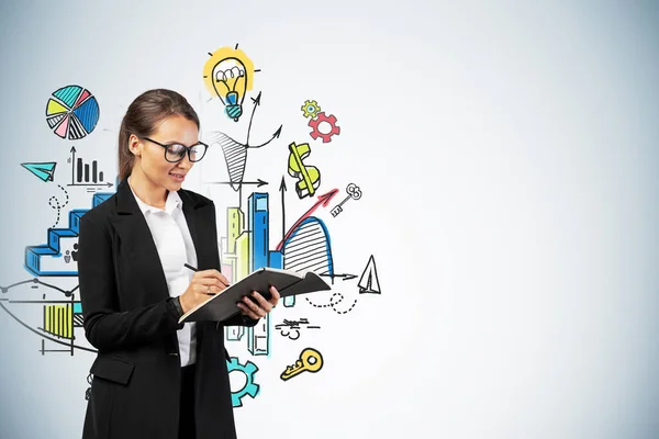 Smiling young European businesswoman or college student in glasses writing in her notebook standing near gray wall with colorful business strategy sketch drawn on it. Mock up