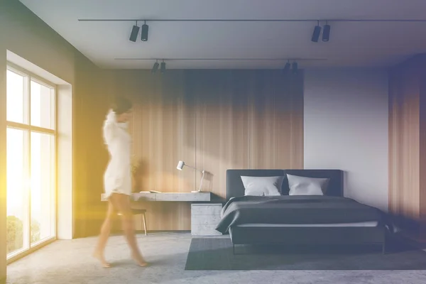 Blurry young woman in nightgown walking in spacious master bedroom with white and wooden walls, concrete floor, comfortable king size bed and table with chair. Toned image
