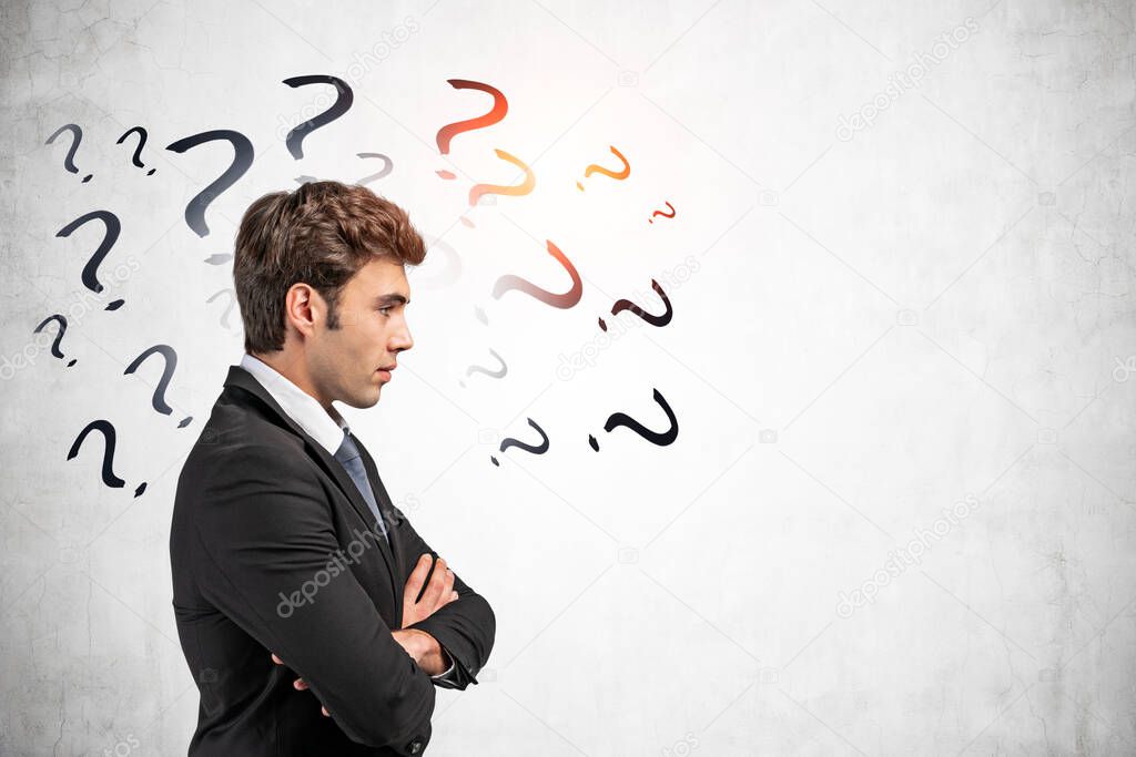 Side view of handsome European businessman standing with crossed arms near concrete wall with question marks written on it. Concept of problem solving and challenge. Mock up