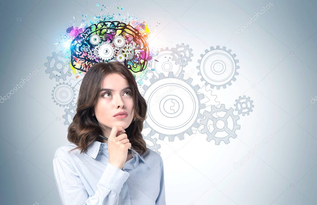Thoughtful young woman with wavy dark hair standing near grey wall with colorful brain with gears drawn on it. Concept of brainstorming and education