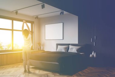 Beyaz ve gri duvarları, ahşap zemini, rahat kral yatağı ve poster çerçevesi olan modern ana yatak odasında duran bulanık, genç bir kadının arka görüntüsü. Tonu ayarlanmış resim