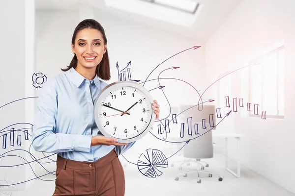 Cheerful young businesswoman with dark hair holding big clock in blurry office with double exposure of financial chart. Concept of time to invest and stock market. Toned image