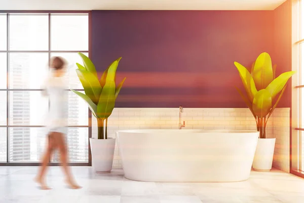 Blurry young woman walking in luxury bathroom with blue and white brick walls, tiled floor and comfortable bathtub with two big potted plants near it. Toned image