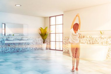 Beyaz mozaik duvarları, fayanslı zemini, rahat bir çift lavabosu ve rahat bir küveti olan güneşli bir banyoda duran genç kadının arka görüntüsü. Bulanık şehir manzaralı bir pencere. Tonu ayarlanmış resim