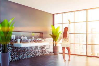 Güneşli panoramik banyoda gri mozaik duvarlar, beton zemin, rahat çift lavabo ve bulanık şehir manzaralı pencere olan genç bir kadın. Tonu ayarlanmış resim