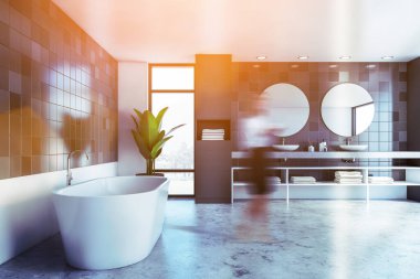 Geniş banyoda siyah fayanslı duvarları, rahat küveti ve yuvarlak aynaları olan çift lavabosu olan bulanık genç bir kadın. Tonu ayarlanmış resim