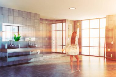 Geniş, güneşli bir banyoda, gri ahşap duvarları, çift lavabosu, duş kabini ve güzellik ürünleriyle dolu rafları olan genç bir kadın. Tonu ayarlanmış resim