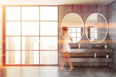 Geniş güneşli banyoda gri ahşap duvarları olan bulanık genç bir kadın oval aynaları olan çift lavabo ve şehir manzaralı bir pencere. Tonu ayarlanmış resim