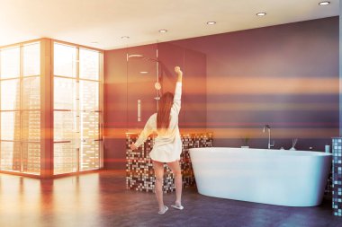 Güneşli banyoda, gri mozaik duvarları, beton zemini, rahat küveti ve cam duş kabini olan genç bir kadının dikiz görüntüsü. Tonu ayarlanmış resim