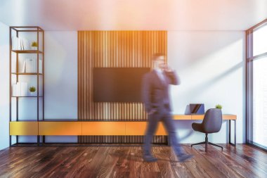 Beyaz ve ahşap duvarları, turuncu bilgisayar masası, kitaplığı ve televizyonu olan bulanık, genç bir işadamı. Tonu ayarlanmış resim