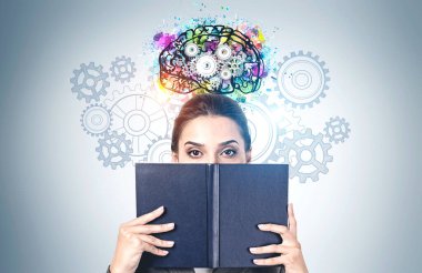 Yüzünü siyah kitapla örten genç bir kadının portresi gri duvarın yanında üzerinde vites çizilmiş renkli beyin çizimiyle duruyor. Eğitim ve beyin fırtınası kavramı