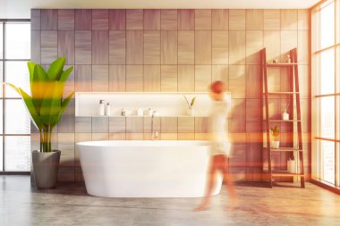 Geniş güneşli banyoda gri ahşap duvarları, rahat küveti ve güzellik ürünleriyle dolu rafları olan bulanık genç bir kadın. Tonu ayarlanmış resim