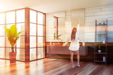 Panoramik banyoda tahta ve gri tuğlalı duvarları olan genç bir kadın ayna ile rahat bir çift lavabo ve güzellik ürünleriyle dolu raflar. Bulanık şehir manzarası. Tonu ayarlanmış resim