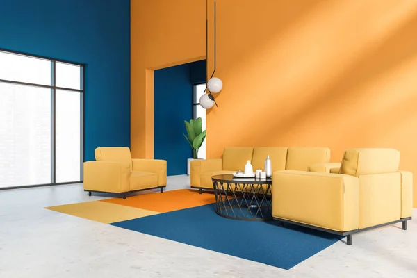 Corner of stylish living room with orange and blue walls, concrete floor, comfortable orange sofa and armchairs near round coffee table and big windows with blurry cityscape. 3d rendering