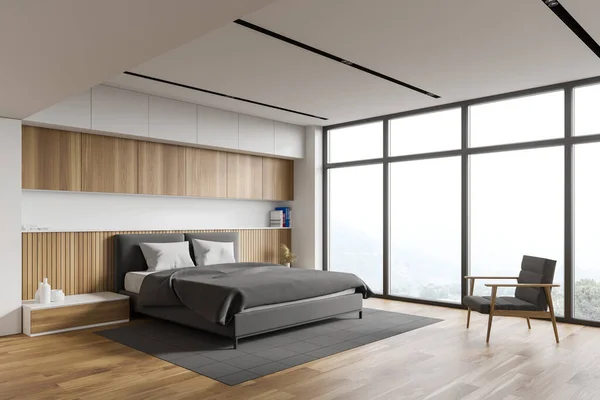 Corner of comfortable master bedroom with white and wooden walls, wooden floor, king size bed standing on grey carpet and armchair near window with blurry mountain view. 3d rendering