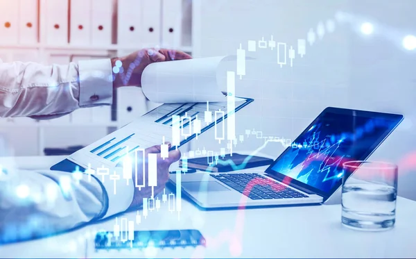 Hands of businessman reading financial report at blurry table with laptop with double exposure of blurry digital graphs. Concept of consulting and stock market. Toned image