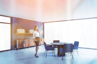 Sarışın iş kadını panoramik ev kütüphanesinde mavi duvarları, beton zemini, mavi sandalyeli yuvarlak masası ve kitaplığı olan akıllı telefonuyla konuşuyor. Bulanık şehir manzarası. Tonu ayarlanmış resim