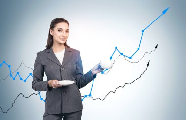 Neşeli genç Avrupalı kadın gri duvarın yanında kahve içiyor ve üzerine grafikler çizilmiş. Yatırım ve iş başarısı kavramı