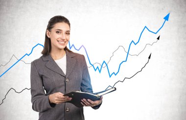 Neşeli genç kadın tüccar elinde defteriyle beton duvarın yanında duruyor ve grafikler büyüyor. Yatırım ve iş başarısı kavramı