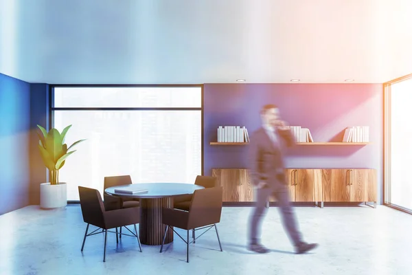 Blurry young man walking and talking on smartphone in home library with blue walls, concrete floor, round table with brown chairs and bookshelves. Toned image