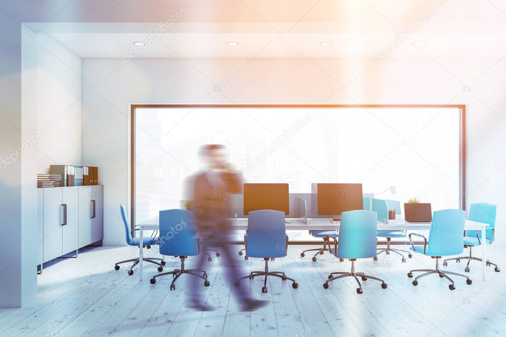 Blurry young businessman walking in panoramic open space office with white walls, long computer table with blue chairs and modern cityscape. Toned image