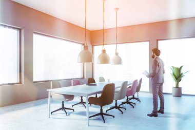 Belgeleri şık minimalist toplantı odasında bej duvarları, beton zemini, pastel sandalyeli uzun konferans masası ve endüstriyel lambaları olan kendine güvenen genç işadamı. Tonu ayarlanmış resim