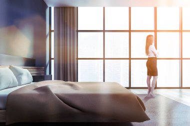 Sarışın genç bir kadın panoramik yatak odasında duruyor. İç kısmı gri duvarlar, ahşap zemin, rahat büyük boy yatak, kitaplık ve bulanık şehir manzarası. Tonu ayarlanmış resim