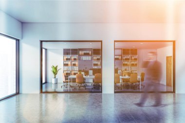 Bulanık genç işadamı, beyaz duvarlı, fayanslı, bilgisayarlı ve turuncu sandalyeli modern açık uzay ofisinde yürüyor. Klasörlü kitaplık. Tonu ayarlanmış resim