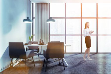 Süslü panoramik yemek odasında, mavi duvarları, halı döşeli, rahat masası ve gri koltukları olan düşünceli genç bir kadın. Tonu ayarlanmış resim