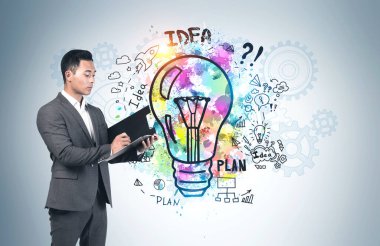 Ciddi bir Asyalı genç işadamı gri duvarın yanındaki panoya renkli bir iş fikri çizimi çizmiş. Yaratıcılık ve planlama kavramı
