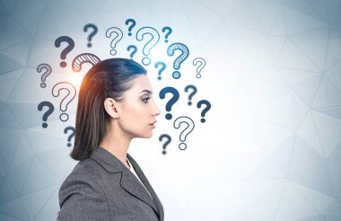Üzerinde soru işaretleri olan gri duvarın yanında duran koyu saçlı, konsantre olmuş genç bir iş kadını. Karar verme ve seçim yapma kavramı. Model