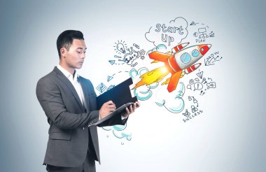 Ciddi bir Asyalı genç işadamı, gri duvarın yanında renkli başlangıç çizimi olan bir panoya yazı yazıyor. Yeni proje başlatma ve iş başarısı kavramı