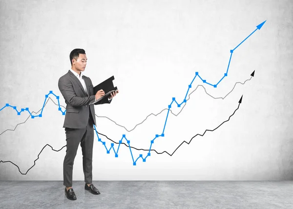 Serious Young Asian Businessman Writing Clipboard Concrete Wall Growing Graphs — Stock Photo, Image