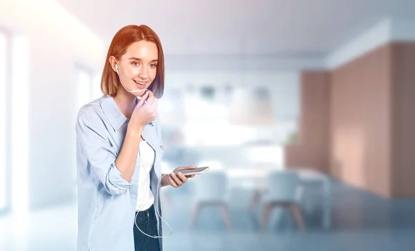 Portrait of smiling European teen girl in casual clothes with headphones talking on smartphone in blurry kitchen. Concept of communication