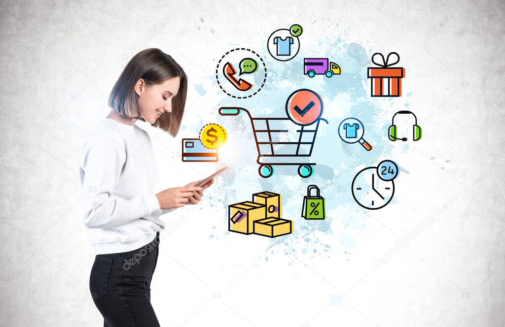 Side view of cheerful young woman with tablet computer standing near concrete wall with colorful online shopping sketch drawn on it. Concept of e commerce.