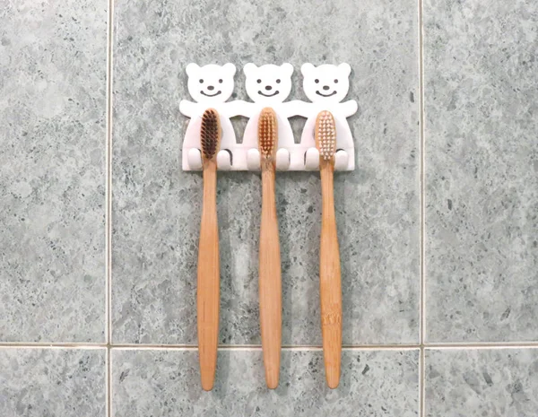 Un conjunto familiar de tres cepillos de dientes de bambú cuelgan en la pared de los baños en soporte de cepillo de dientes. Estilo de vida ecológico . — Foto de Stock