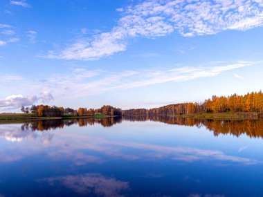 Sakin sulara yansıyan ağaçlarla ve bulutlarla dolu bir manzara.