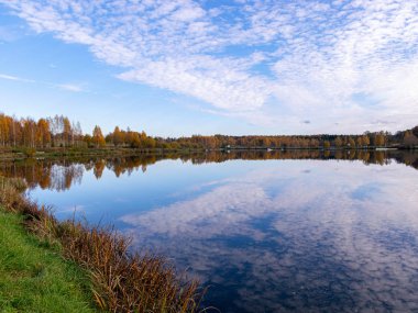 Sakin sulara yansıyan ağaçlarla ve bulutlarla dolu bir manzara.