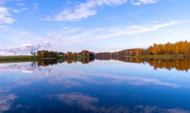 Sakin sulara yansıyan ağaçlarla ve bulutlarla dolu bir manzara.