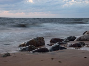 Kayalık bir sahilde gün batımı manzarası, ön planda bulanık dalgalar ve kayalar.