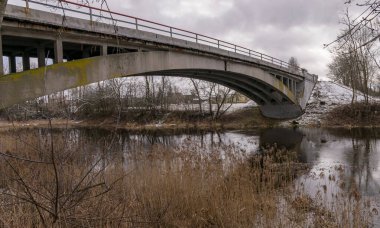 Çalılar, kurumuş dallar ve eski otlarla kaplanmış küçük ve vahşi bir nehir üzerindeki beton kemerli köprü.