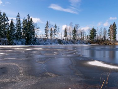 Güneşli bir kış manzarası, ön planda donmuş bir gölün dokusu, buzdaki gökyüzünün parlaklığı.