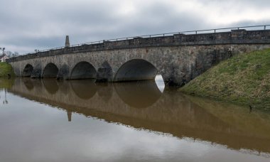 İlkbaharda taşan nehir manzarası, nehir üzerinde kemerli taş köprü, suda ayna yansıması.