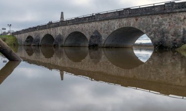 İlkbaharda taşan nehir manzarası, nehir üzerinde kemerli taş köprü, suda ayna yansıması.