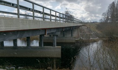 Manzarada kar, nehrin üzerindeki beton köprü, bahar günü, çıplak çalılar ve eski otlar.