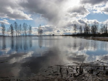Göllü güneşli bir manzara, suda parlayan bulutlar, uzaktaki çıplak ağaç siluetleri.